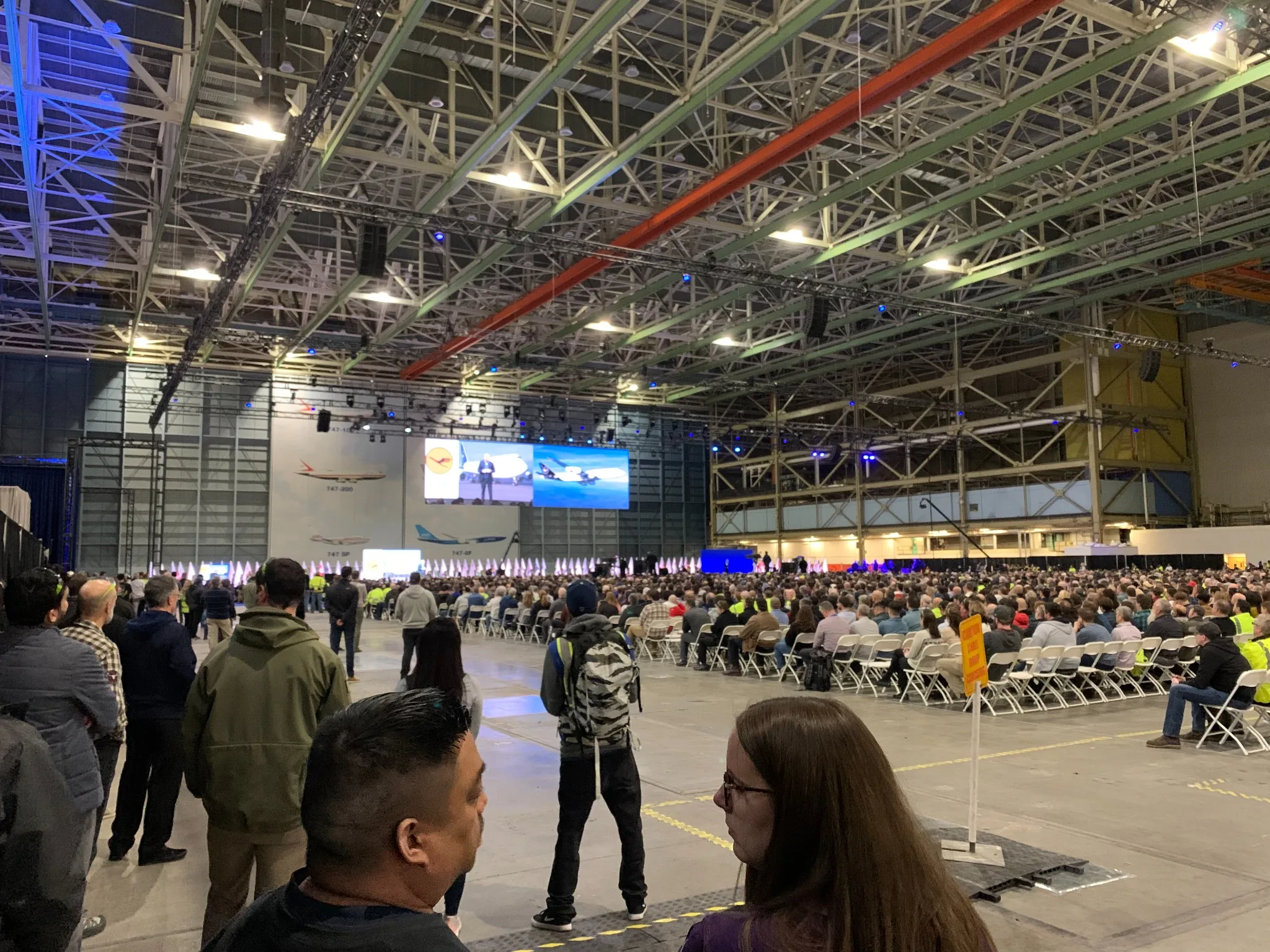 Large-scale live event inside an industrial venue with a seated audience, overhead rigging and stage presentation
